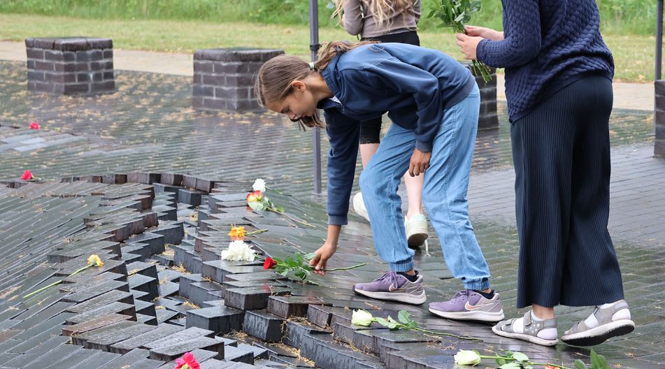 Kinder legen Blumen auf ein dunkles Gedenkmal im Freien nieder.