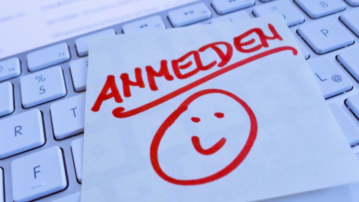 Ein handgeschriebener Zettel mit dem Wort 'Anmelden' und einem lächelnden Gesicht liegt auf einer Computertastatur. Ein handgeschriebener Zettel mit dem Wort 'Anmelden' und einem lächelnden Gesicht liegt auf einer Computertastatur.