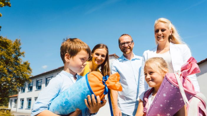 Zwei Kinder mit bunten Schultüten stehen vor einer Schule, begleitet von drei lächelnden Erwachsenen bei strahlendem Sonnenschein. Zwei Kinder mit bunten Schultüten stehen vor einer Schule, begleitet von drei lächelnden Erwachsenen bei strahlendem Sonnenschein.